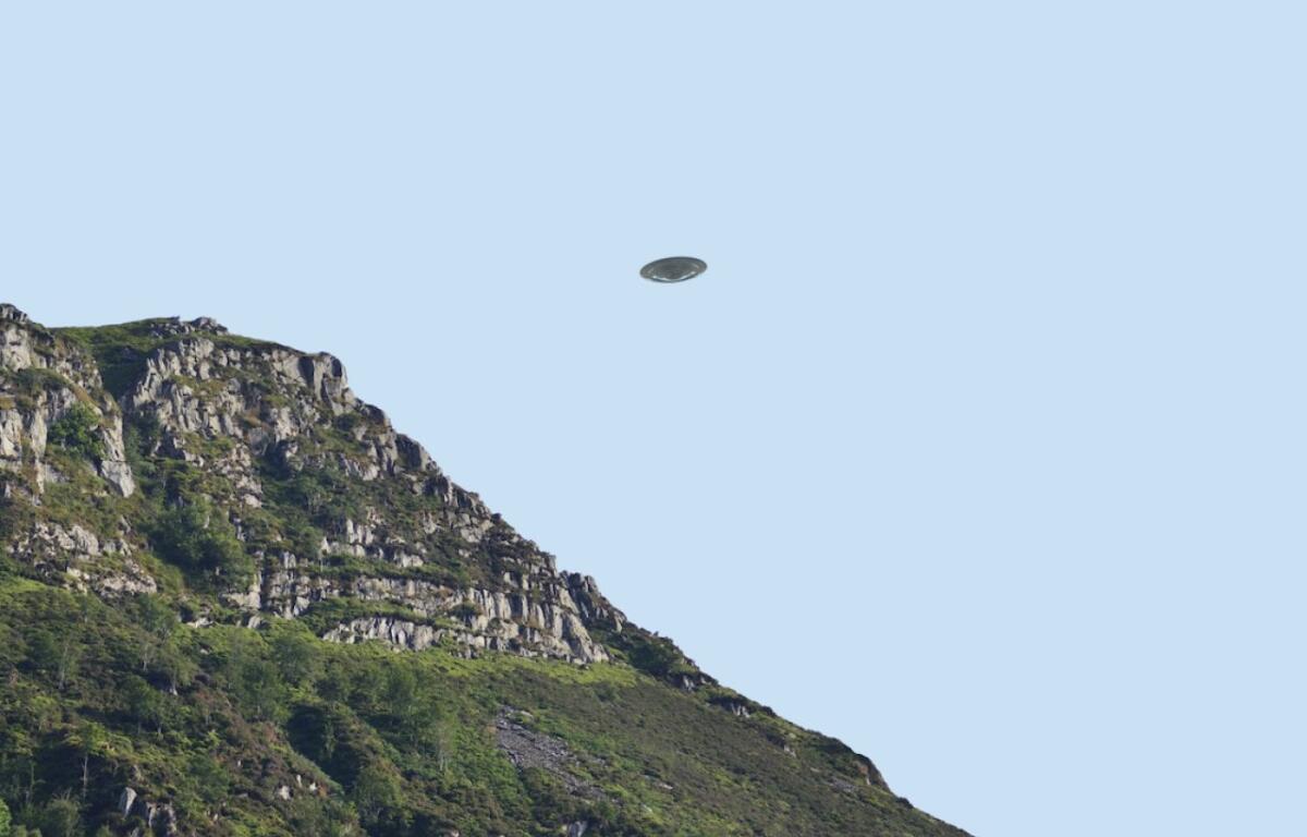 Rocky hillside with green vegetation on the left, under a clear blue sky, and a small flying saucer-shaped craft hovering in the center.