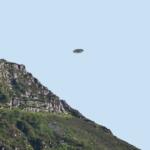 Rocky hillside with green vegetation on the left, under a clear blue sky, and a small flying saucer-shaped craft hovering in the center.