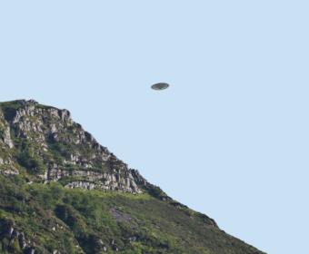 Rocky hillside with green vegetation on the left, under a clear blue sky, and a small flying saucer-shaped craft hovering in the center.