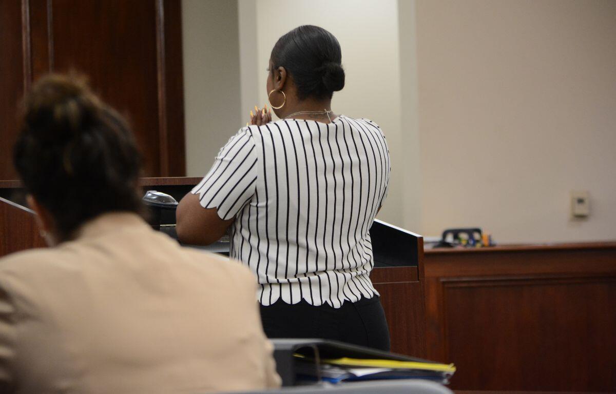 Woman standing at a wooden podium in a courtroom/meeting room, seen from behind, wearing a white blouse with black vertical stripes and a bun, with another person seated in the foreground.