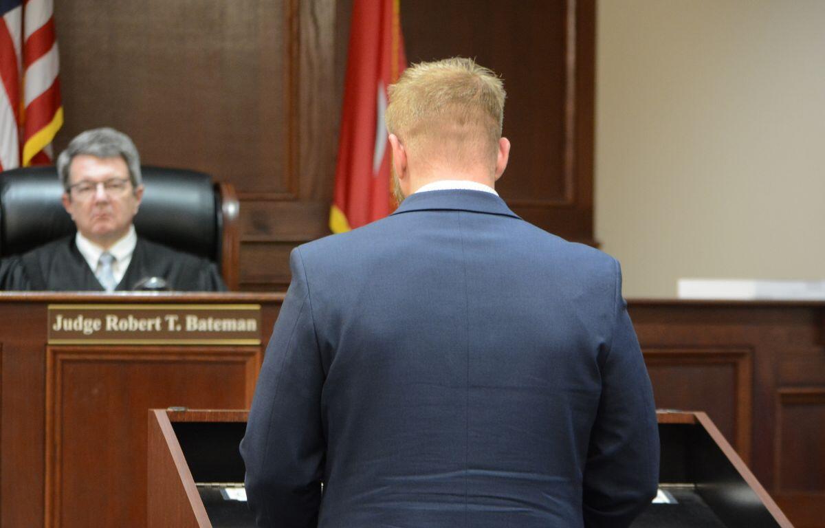 Man in a blue suit stands at a courtroom lectern, facing a judge seated behind a bench labeled 'Judge Robert T. Bateman'