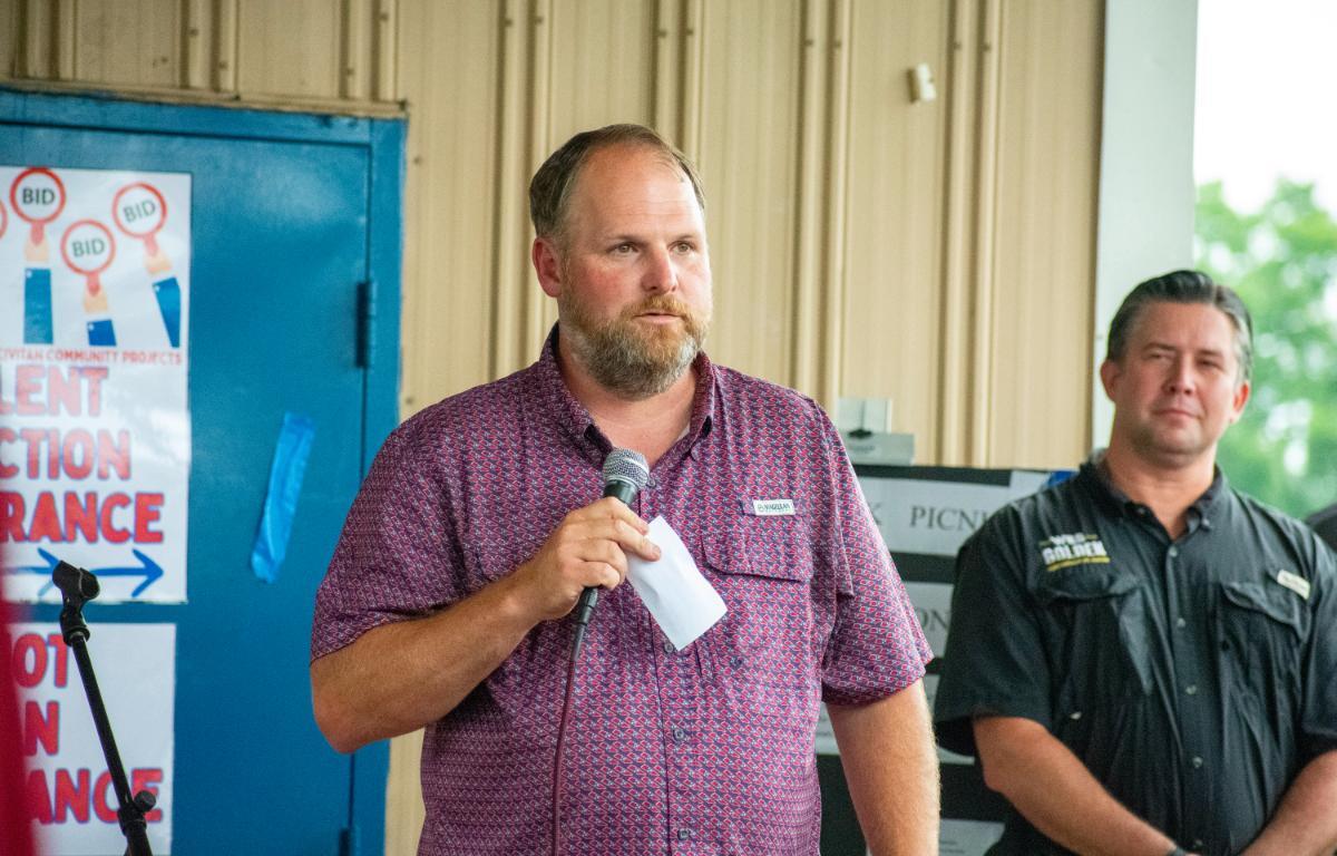 Man in a maroon patterned shirt speaks into a handheld microphone at a community event, holding a folded note. Behind him is a blue wall with posters.