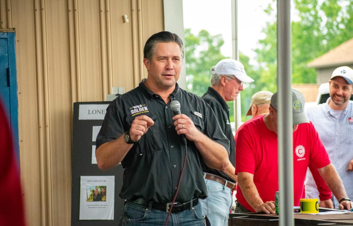 Man in a black shirt speaks into a handheld microphone at an outdoor event, with several people and a table in the background.