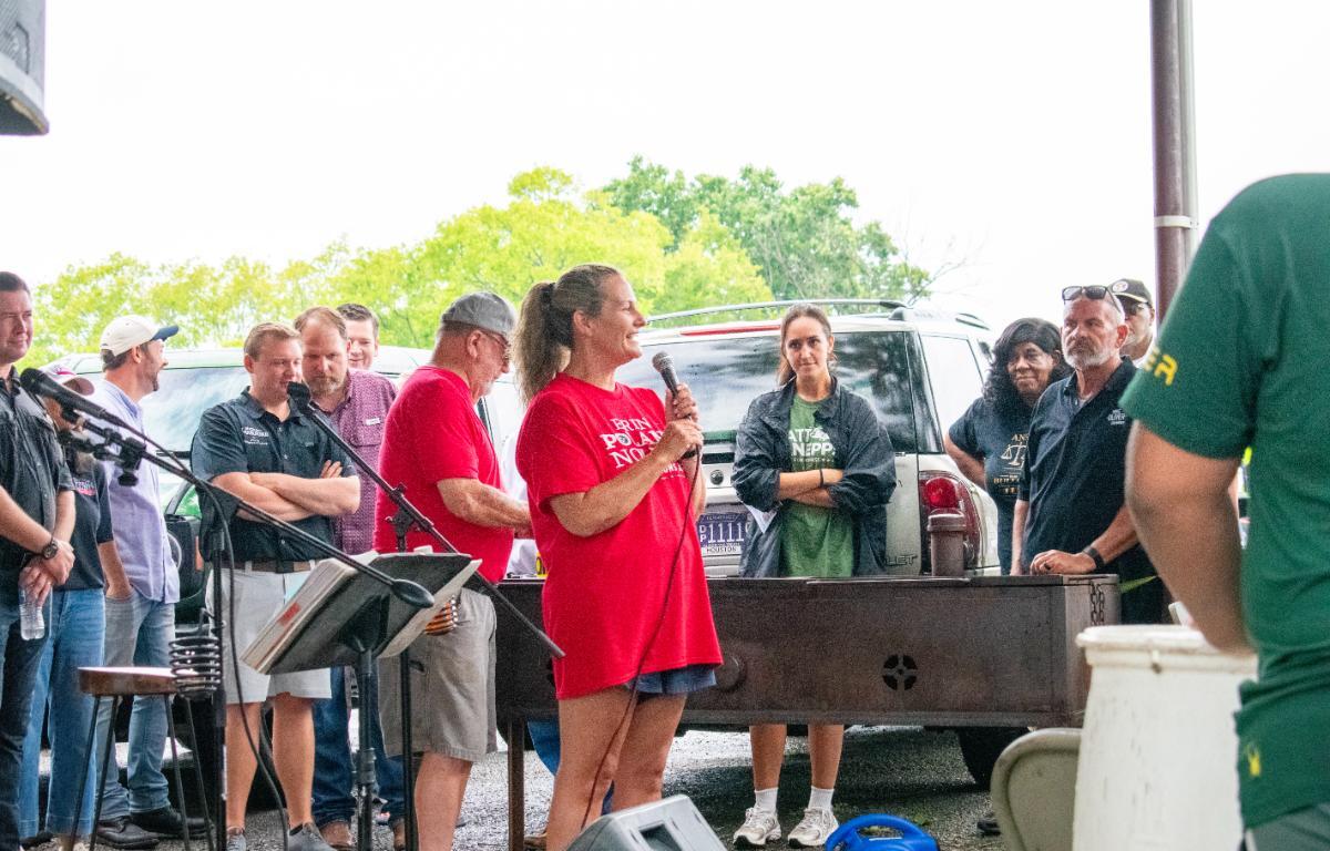 Woman in a red shirt speaks into a microphone at an outdoor fundraiser, with a crowd gathered around a grill and a pickup in the background.