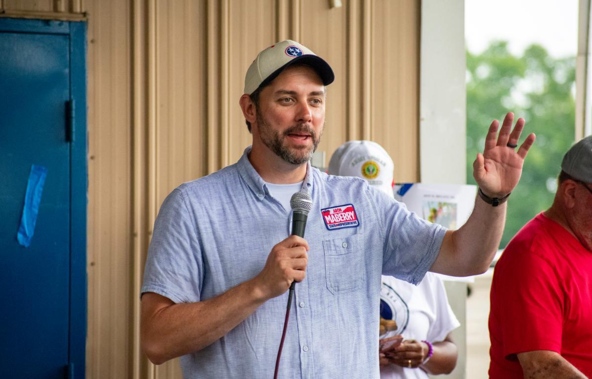 Man in a gray cap and light blue button-down shirt speaks into a handheld microphone, gesturing with his raised right hand.