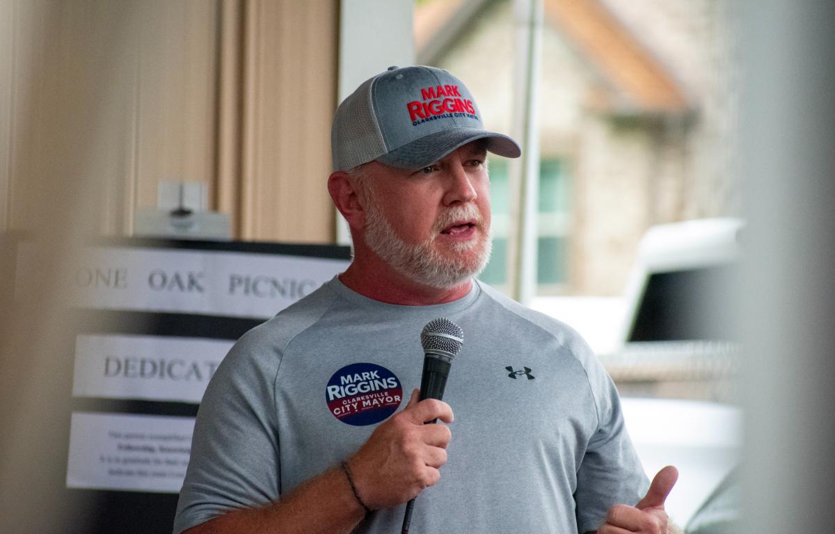Man in a gray Under Armour shirt and cap speaking into a handheld microphone at a memorial or community event, with a Mark Riggins badge visible on his chest.