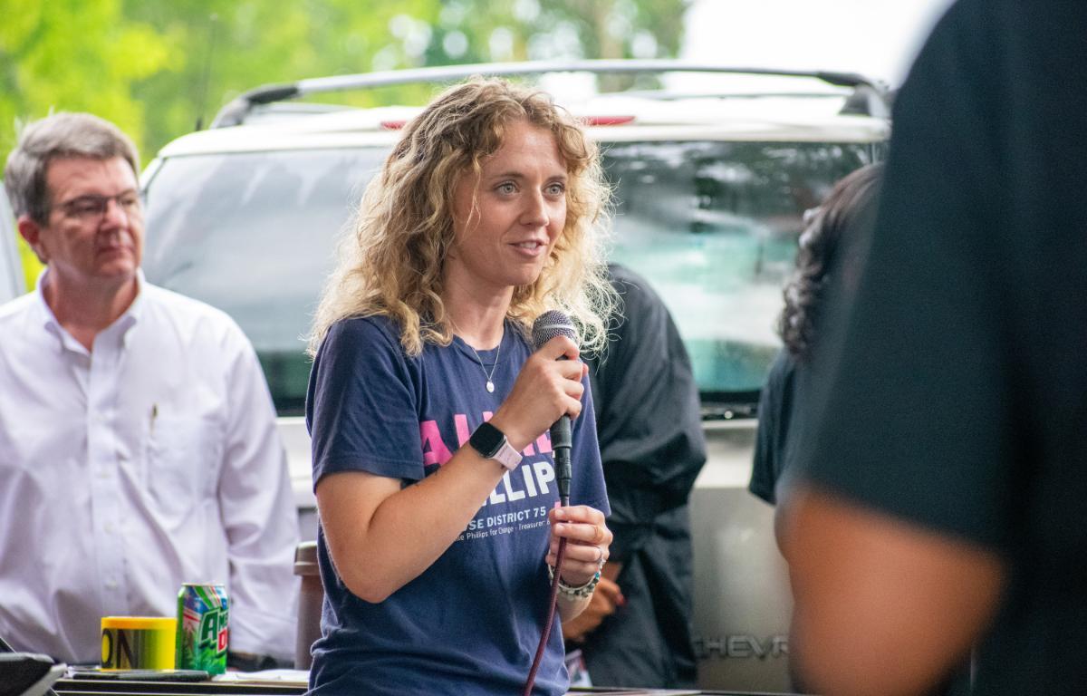 Woman with curly blonde hair speaks into a microphone at an outdoor gathering, wearing a navy T-shirt and smartwatch.