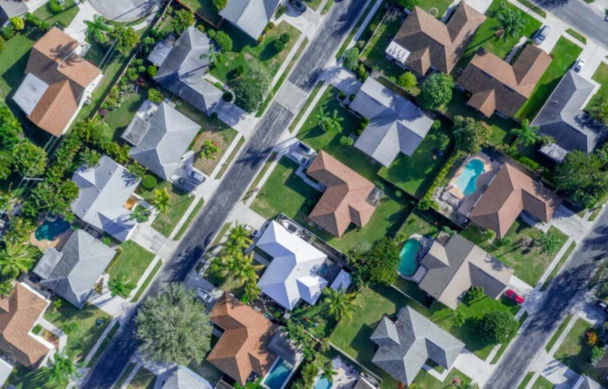 Aerial view of a suburban neighborhood with houses, green lawns, and several backyard pools visible.