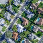 Aerial view of a suburban neighborhood with houses, green lawns, and several backyard pools visible.