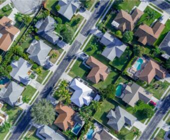 Aerial view of a suburban neighborhood with houses, green lawns, and several backyard pools visible.