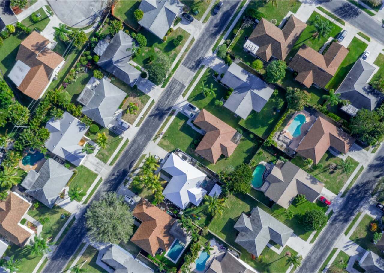 Aerial view of a suburban neighborhood with houses, green lawns, and several backyard pools visible.