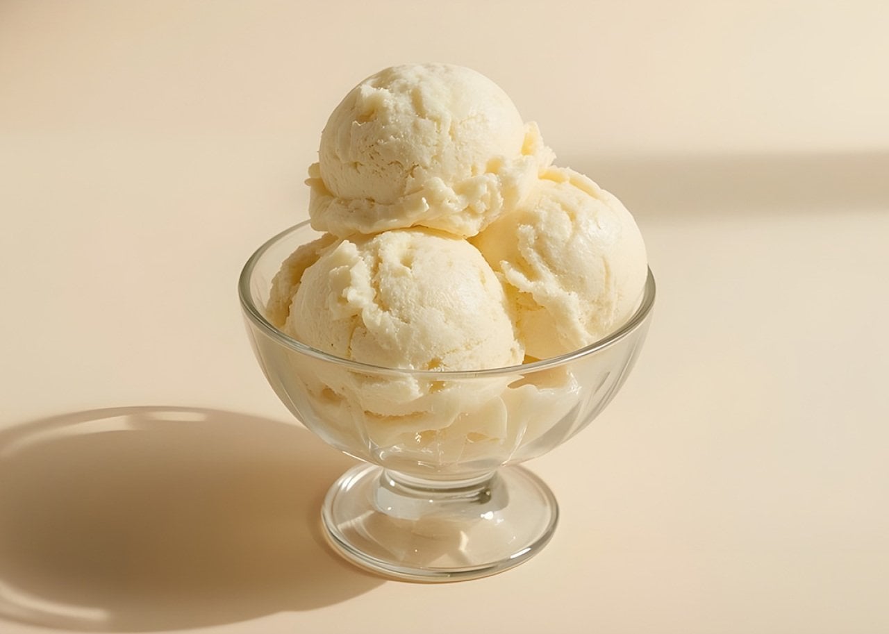 Vanilla ice cream scoops stacked in a clear glass dessert bowl on a pale background, with a soft shadow nearby.