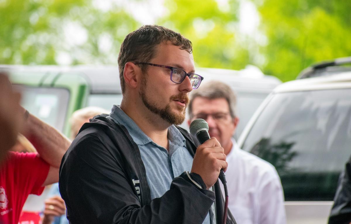 Man wearing glasses speaks into a handheld microphone at an outdoor event, with others and vehicles in a blurred background.