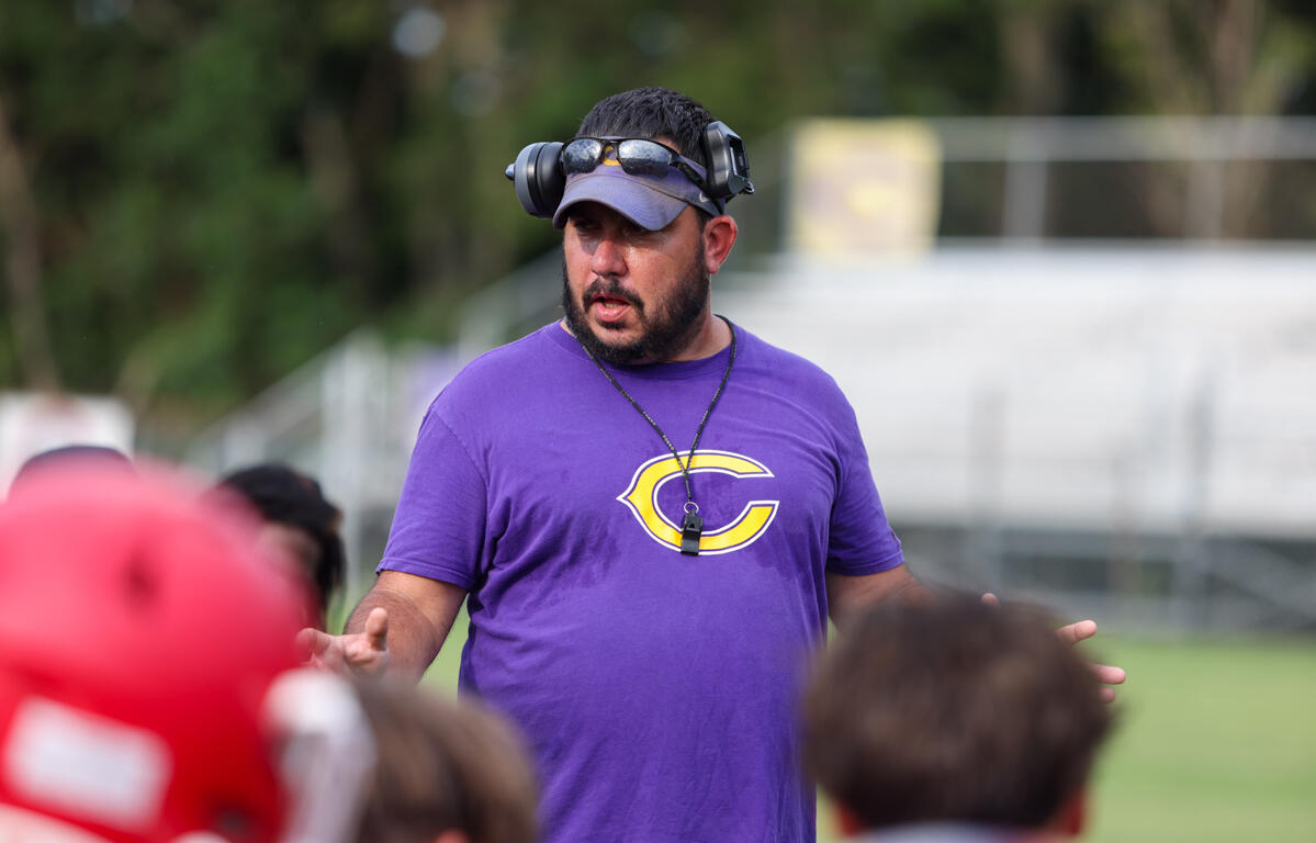Coach wearing a purple shirt with a yellow 'C' logo speaks to children on a sports field, headset around his head and whistle on a neck strap.