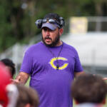 Coach wearing a purple shirt with a yellow 'C' logo speaks to children on a sports field, headset around his head and whistle on a neck strap.