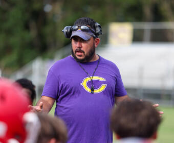 Coach wearing a purple shirt with a yellow 'C' logo speaks to children on a sports field, headset around his head and whistle on a neck strap.