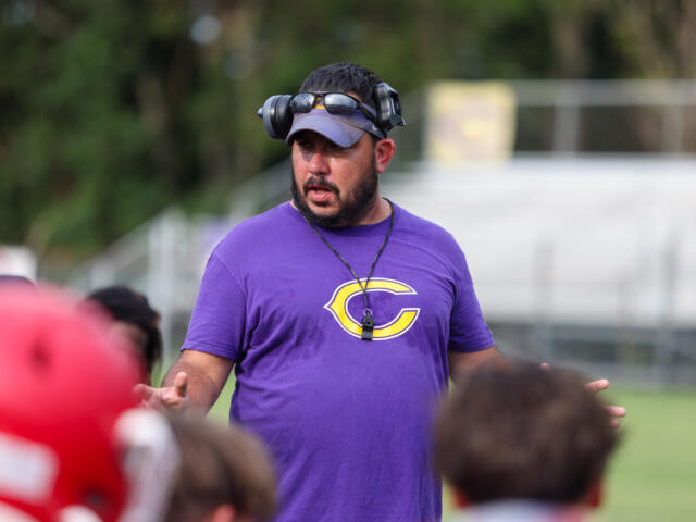 Coach wearing a purple shirt with a yellow 'C' logo speaks to children on a sports field, headset around his head and whistle on a neck strap.