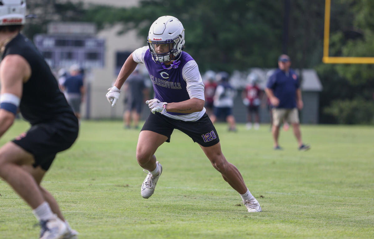Lacrosse player in a purple jersey (#120) sprinting on a green field with a white helmet and gloves, during a game.