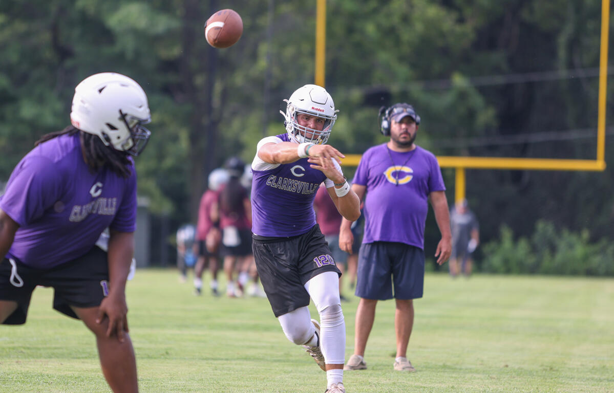 Quarterback in a purple uniform throws a football during practice on a grassy field, with a coach watching in the background.