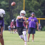 Quarterback in a purple uniform throws a football during practice on a grassy field, with a coach watching in the background.