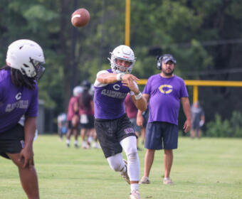 Quarterback in a purple uniform throws a football during practice on a grassy field, with a coach watching in the background.