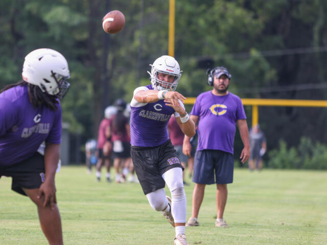 Quarterback in a purple uniform throws a football during practice on a grassy field, with a coach watching in the background.