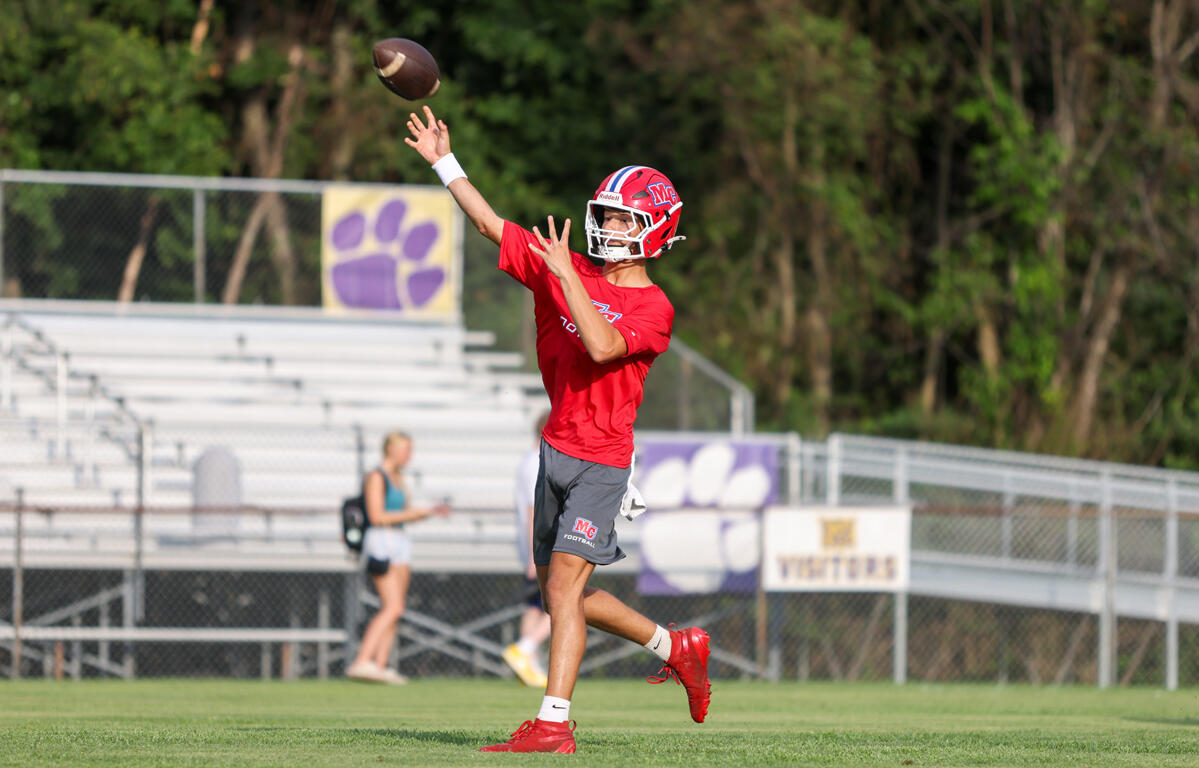 Quarterback in a red jersey and helmet throws a football on a grassy field during practice, with bleachers and a purple paw banner in the background.