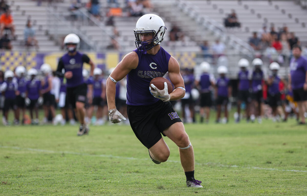 Football player in a purple jersey and white helmet runs with the ball on a grassy field, with teammates and bleachers in the background.