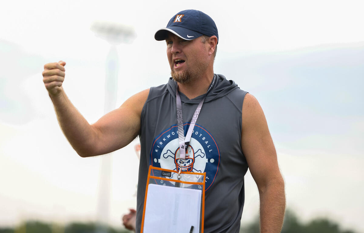 Coach wearing a navy cap and gray sleeveless shirt raises a clenched fist and speaks to players on a sports field during practice, with a clipboard hanging from a lanyard.
