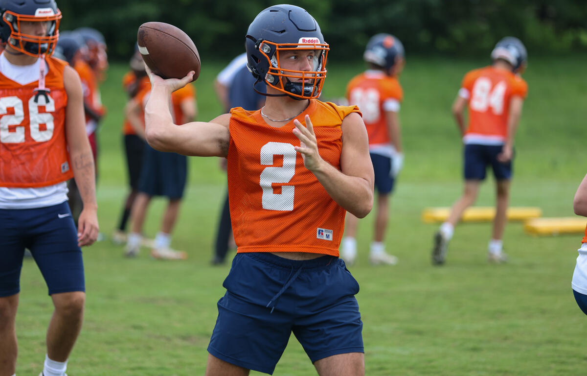 Quarterback in orange practice jersey throws a football during a drill on a green field.2 with helmet and navy shorts visible.