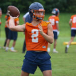 Quarterback in orange practice jersey throws a football during a drill on a green field.2 with helmet and navy shorts visible.