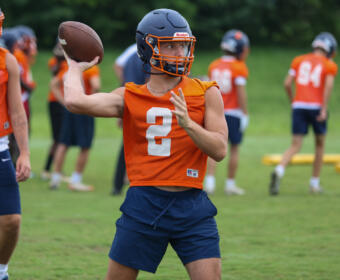 Quarterback in orange practice jersey throws a football during a drill on a green field.2 with helmet and navy shorts visible.