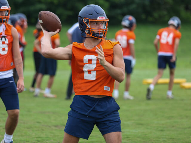 Quarterback in orange practice jersey throws a football during a drill on a green field.2 with helmet and navy shorts visible.