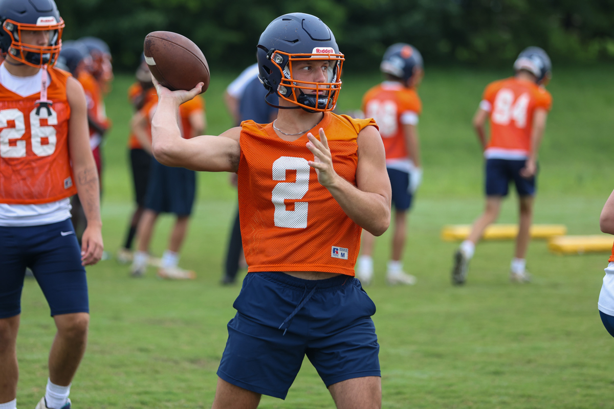 Quarterback in orange practice jersey throws a football during a drill on a green field.2 with helmet and navy shorts visible.