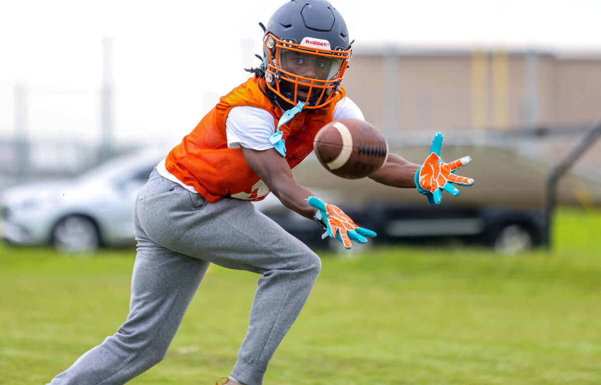 Youth football player in an orange practice vest reaches for a pass, helmet on, teal gloves visible, on a grassy field.