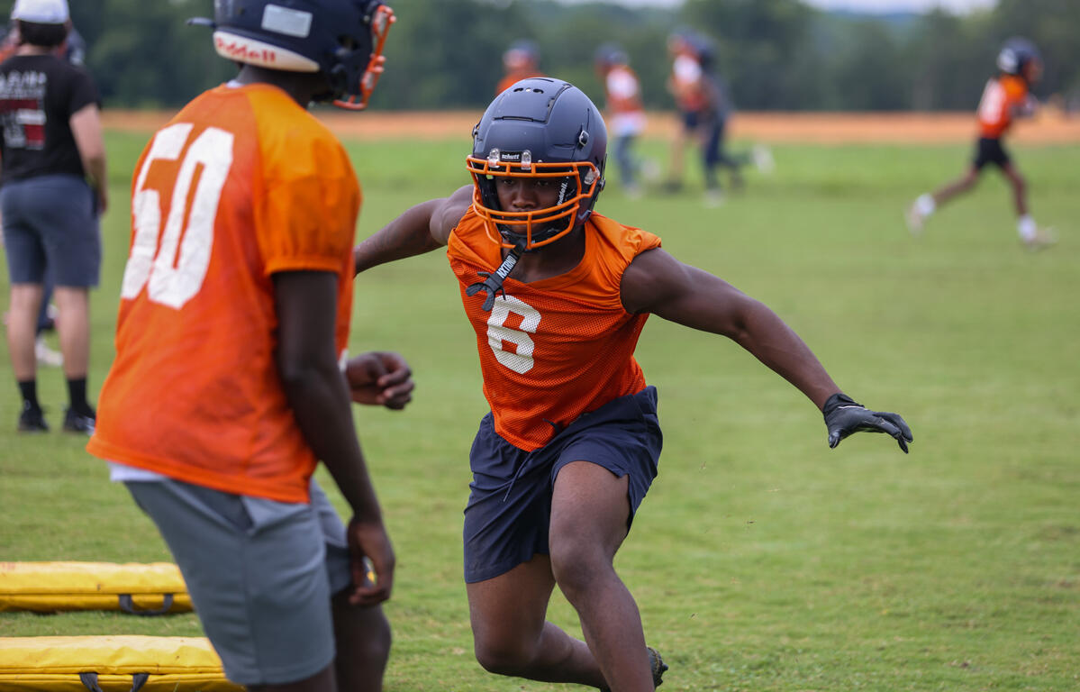 Young football player in an orange jersey and helmet sprints across a grassy field during practice, with a teammate nearby.
