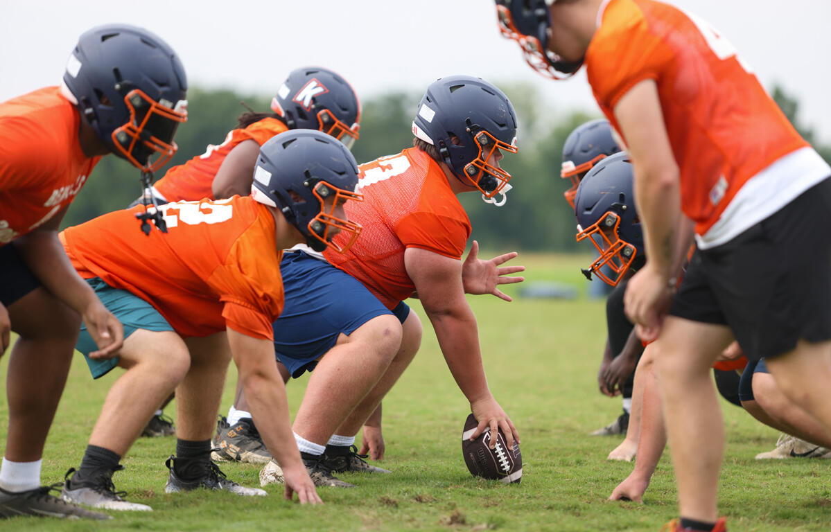 Football players in orange jerseys lined up at the line of scrimmage, center snapping the ball during a practice play.
