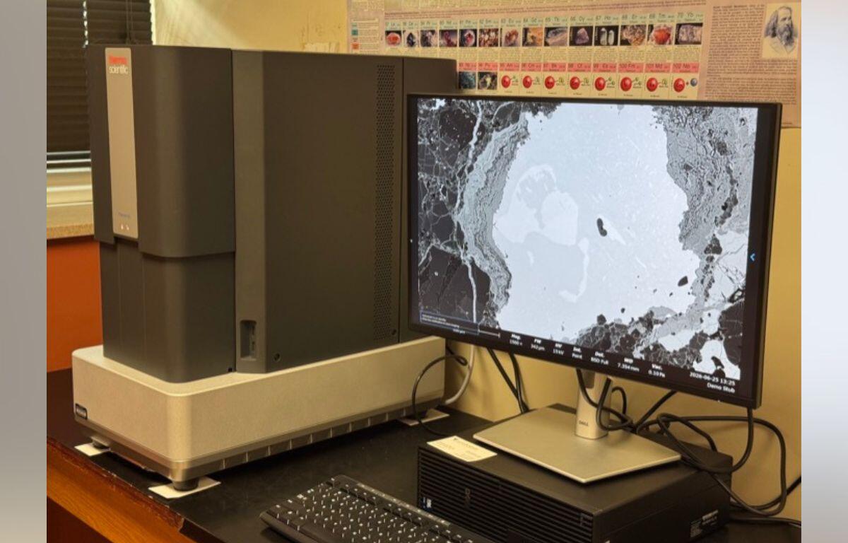 Scanning electron microscope on a lab bench with a monitor displaying a grayscale micrograph of a sample.