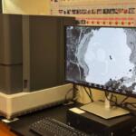 Scanning electron microscope on a lab bench with a monitor displaying a grayscale micrograph of a sample.