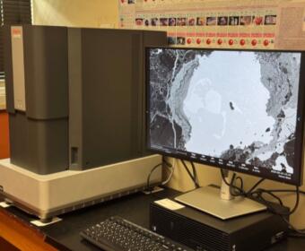 Scanning electron microscope on a lab bench with a monitor displaying a grayscale micrograph of a sample.