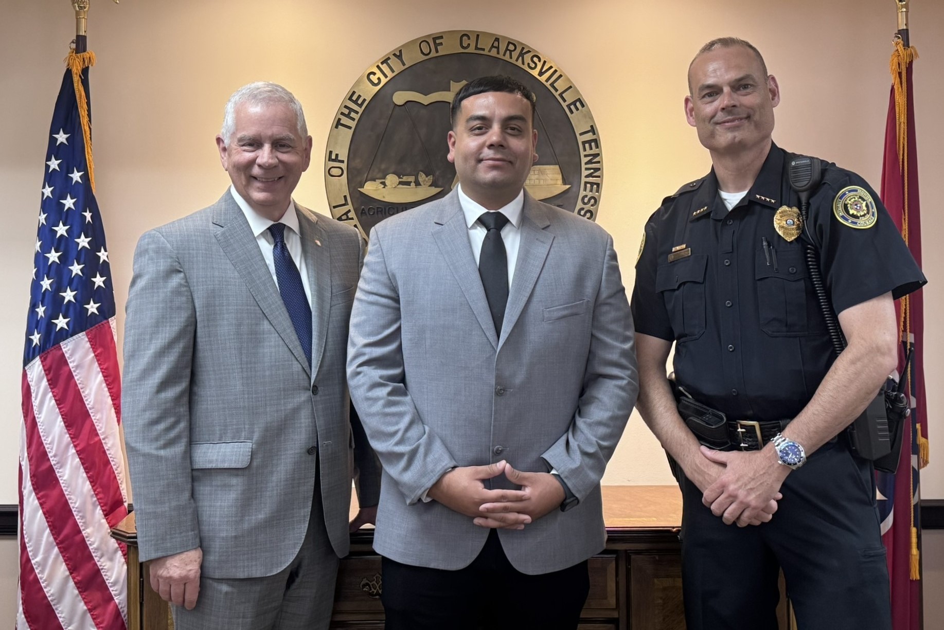 Three men in suits and a police uniform posing in front of the Clarksville city seal with American flags behind them, in a formal portrait. (informative)