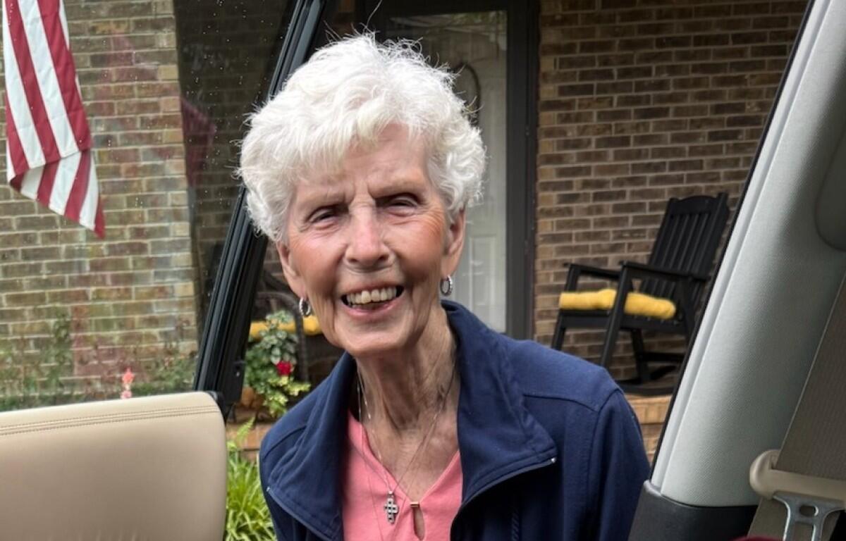 Smiling elderly woman with short white hair leaning into a car door, wearing pink top and navy jacket, outside a brick house.