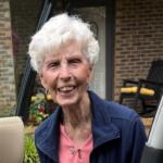 Smiling elderly woman with short white hair leaning into a car door, wearing pink top and navy jacket, outside a brick house.