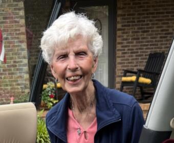 Smiling elderly woman with short white hair leaning into a car door, wearing pink top and navy jacket, outside a brick house.