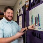 Smiling man in a light blue polo points to a multicolored bar chart posted on a purple hallway bulletin board to explain the data.