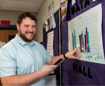 Smiling man in a light blue polo points to a multicolored bar chart posted on a purple hallway bulletin board to explain the data.