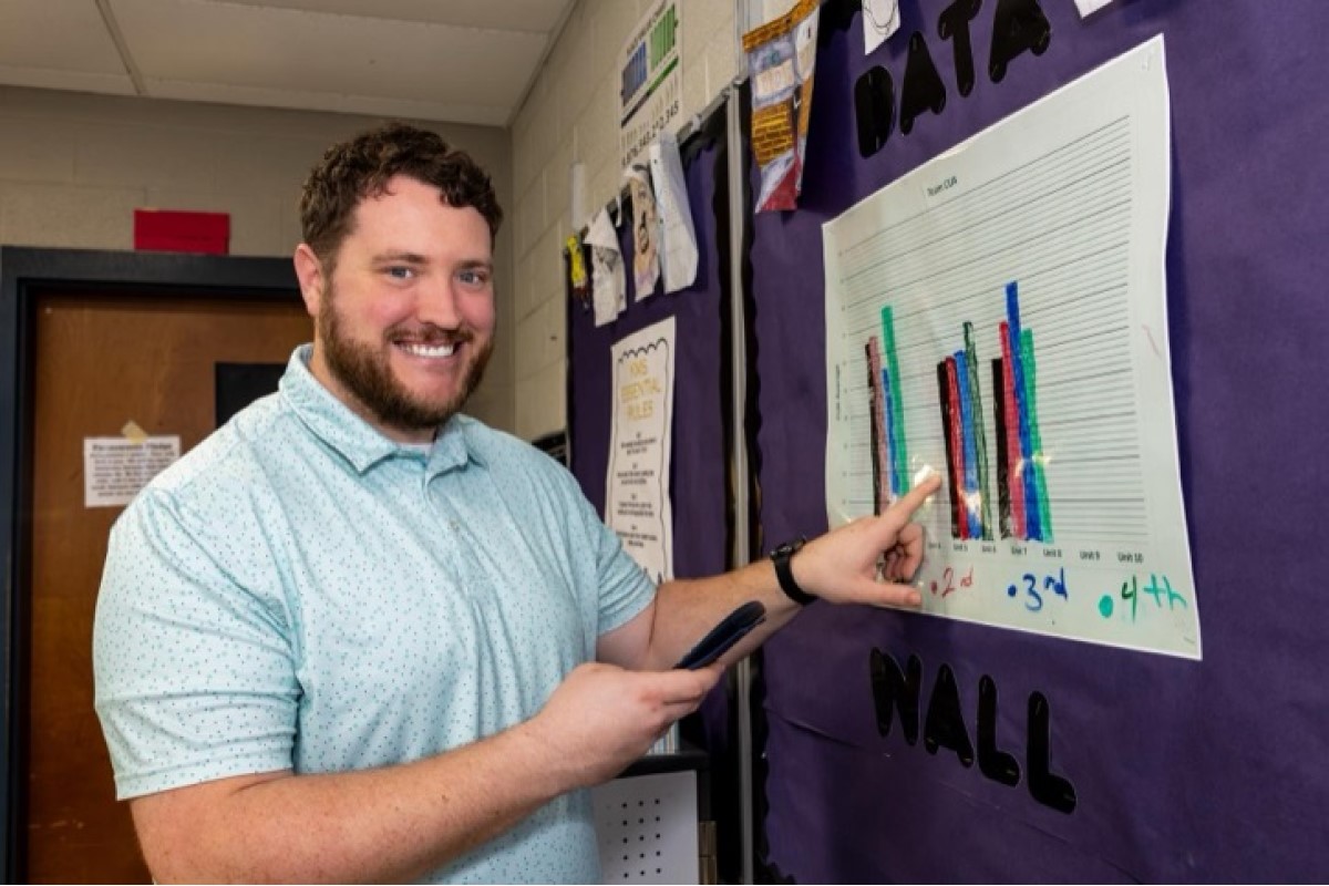Smiling man in a light blue polo points to a multicolored bar chart posted on a purple hallway bulletin board to explain the data.