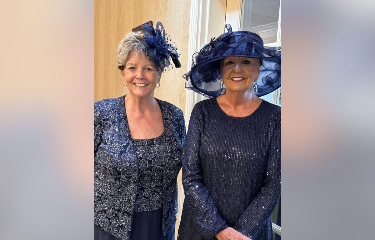 Two women wearing navy outfits and decorative hats smile for the camera indoors.