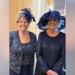 Two women wearing navy outfits and decorative hats smile for the camera indoors.