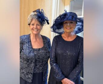 Two women wearing navy outfits and decorative hats smile for the camera indoors.